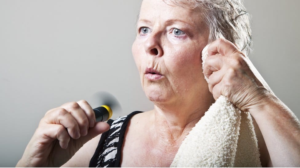 A woman going through a hot flush - she's holding a fan in one hand, while she wipes her face with a towel with the other hand