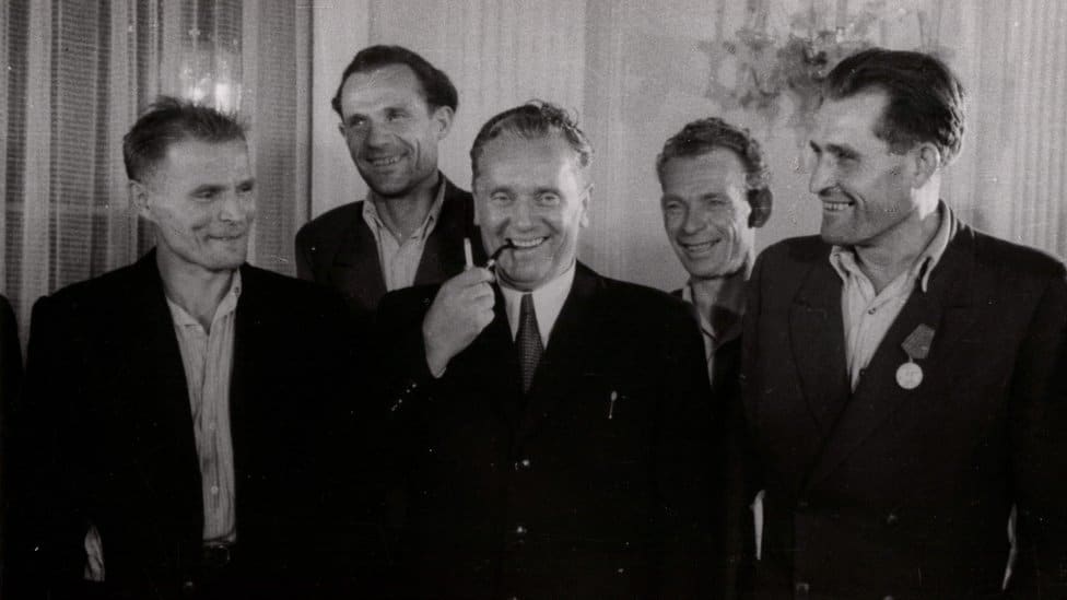 Tito u sredini puši lulu, oko njega okupljeni nasmejani radnici