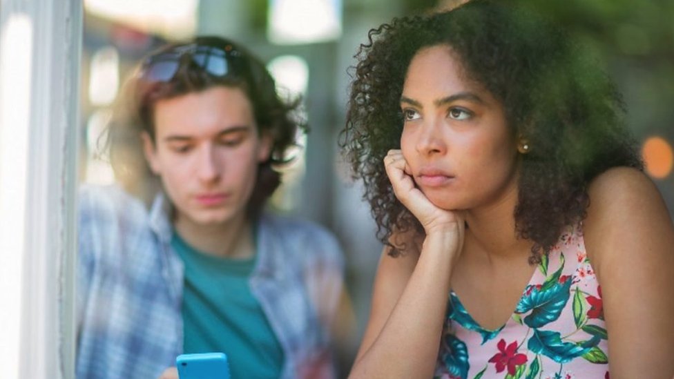 Woman who looks unhappy stares out the window as her partner looks at his phone