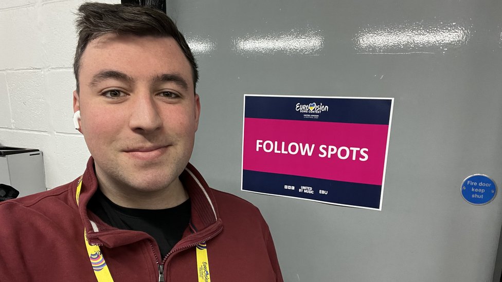 Luke wearing a Eurovision lanyard and standing by a door with a sign saying 'follow spots'