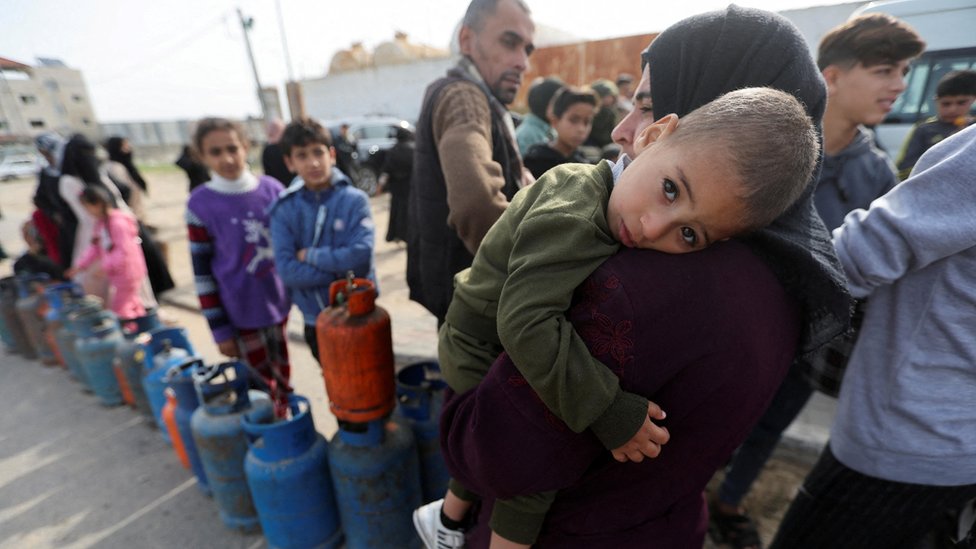 A woman carries a child as Palestinians gather to fill liquid gas cylinders, during a temporary truce between Hamas and Israel, in Rafah in the southern Gaza Strip