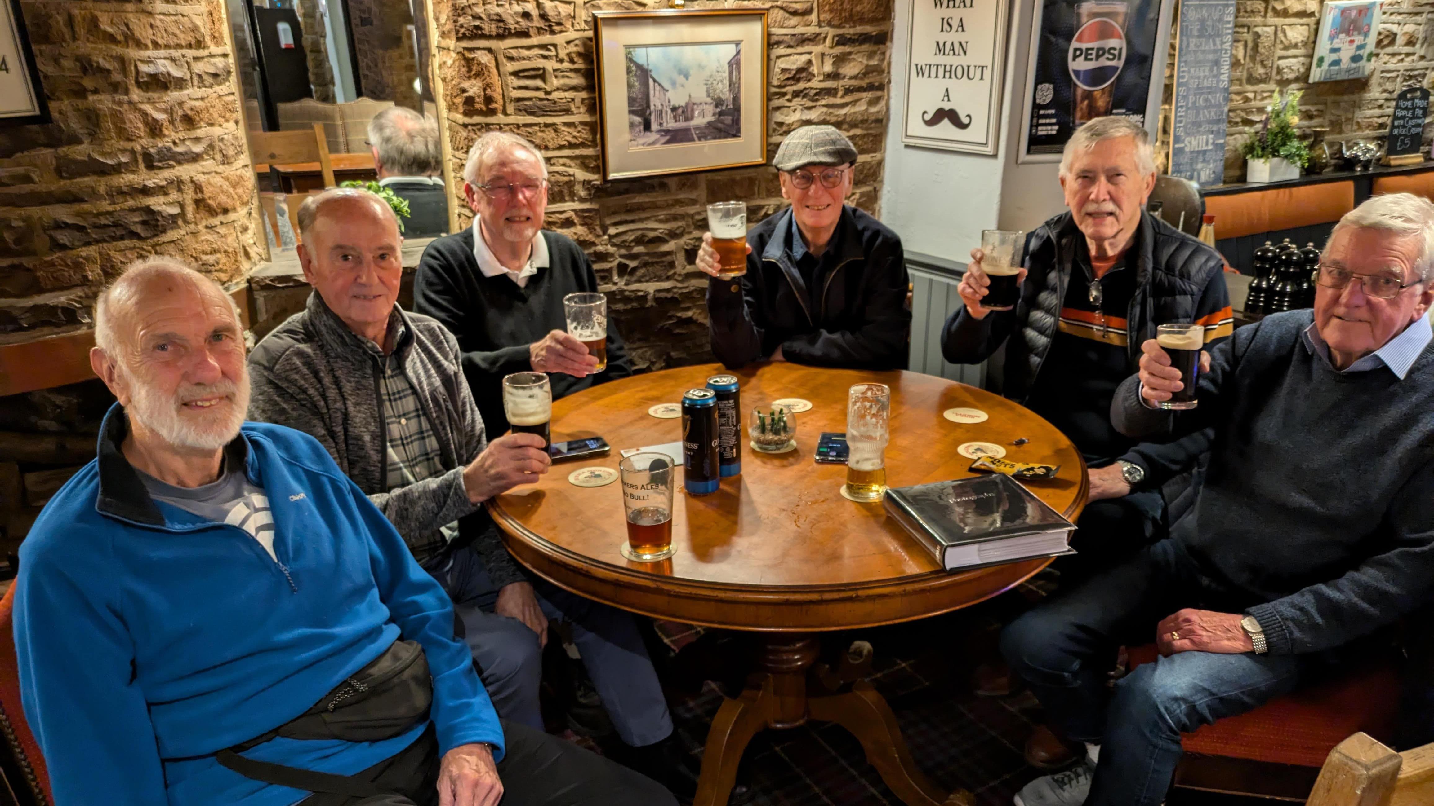 Six gentlemen in their eighties sit around a pub table holding their beers up