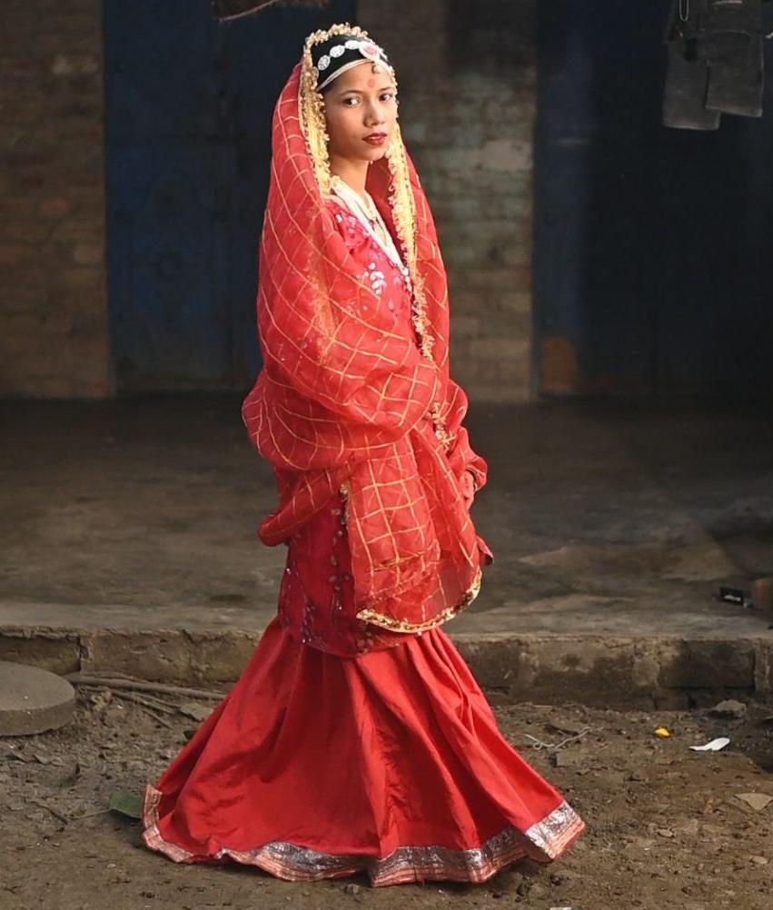 A girl poses for the camera wearing colourful red clothing and jewellery, she's standing outside on a street and looks directly at the camera. The scene is well lit which makes the clothing looks vibrant. 