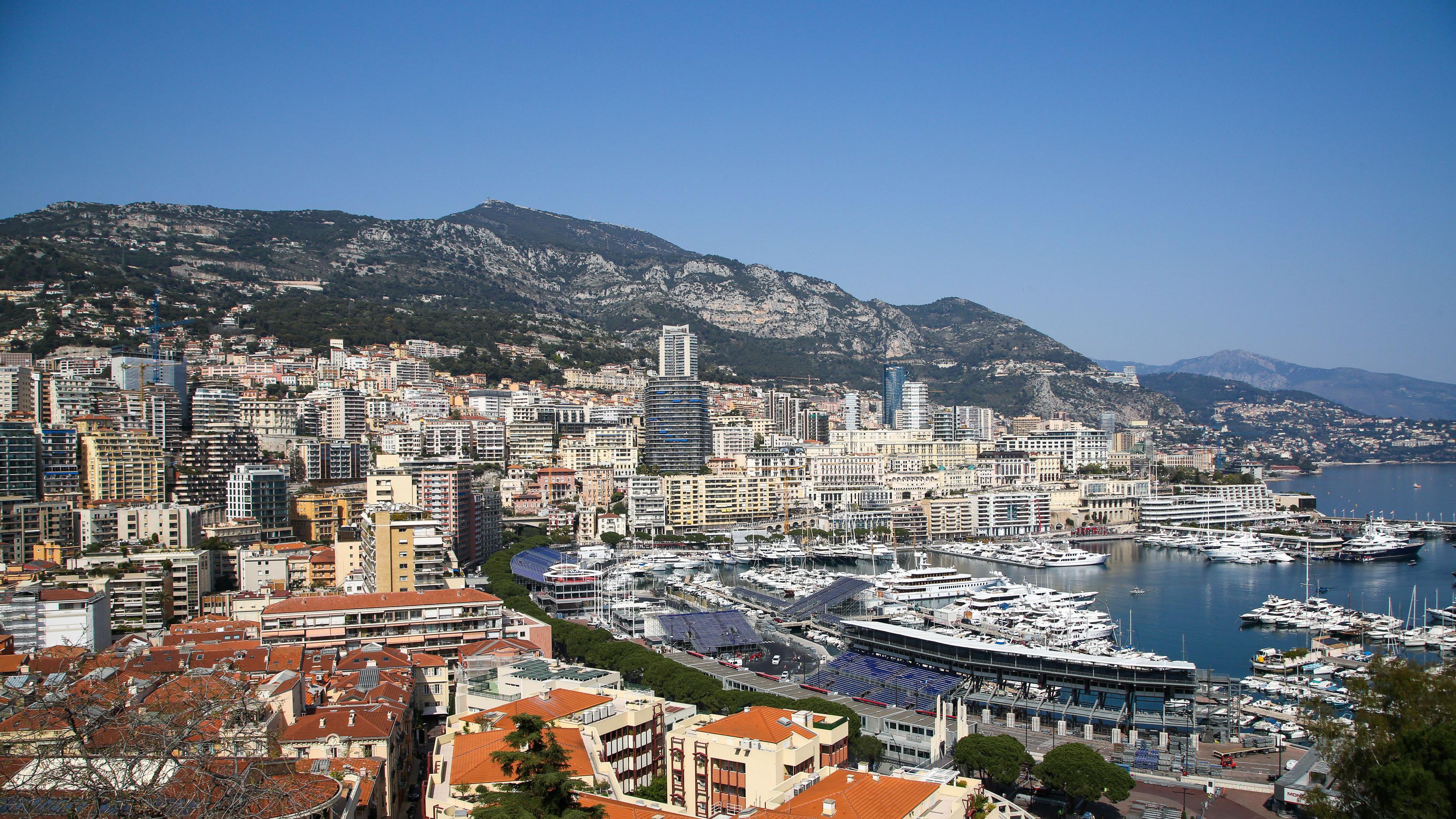 Aerial view of Port Hercules, filled with yachts, and the tower blocks of Monte Carlo in Monaco, with mountains rising behind them and Cap Martin visible in the distance