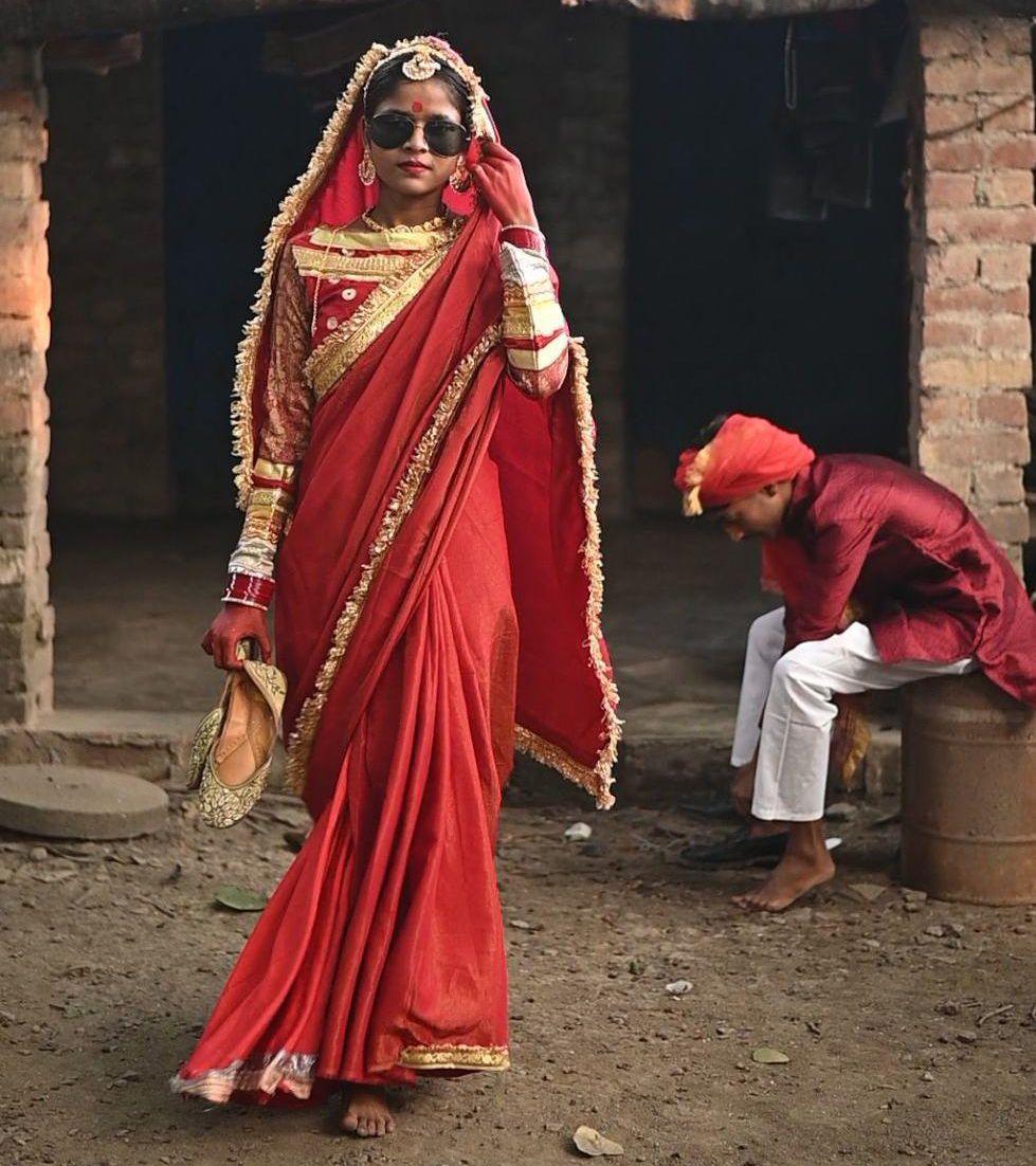 A girl models at a fashion show that has gone viral, she is walking down a street while wearing colourful red clothing, jewellery and sunglasses, and a man is sitting on the side of the street behind her putting his shoes on.