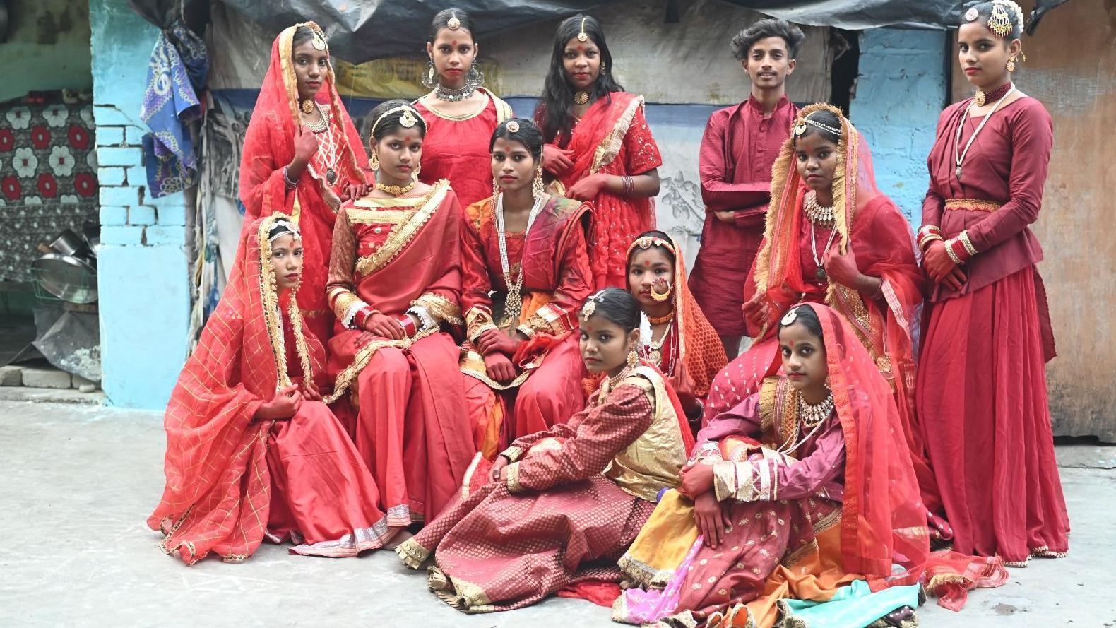 A group of Indian children at a fashion show on a street, all wearing colourful red outfits and ornate jewellery including Maang Tikka, necklaces, earrings and bracelets. 