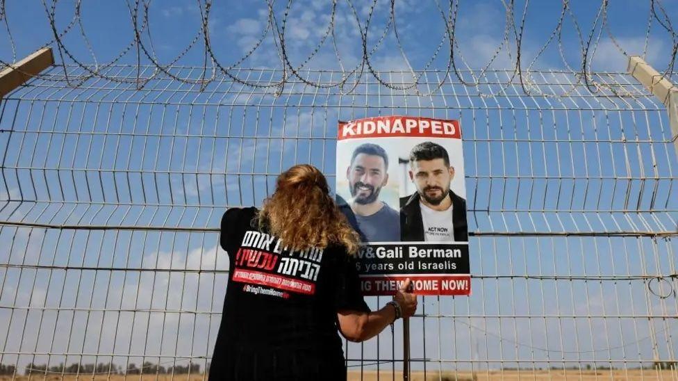 A woman attaches a poster to a fence, showing the faces of two men with the word 'Kidnapped' at the top